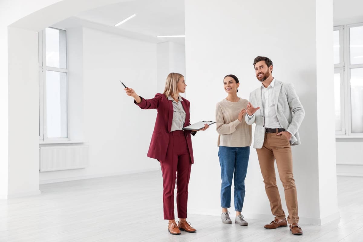 A real estate agent stands together with a couple in an all-white apartment with white flooring and white walls, explaining a detail she is pointing to with her arm