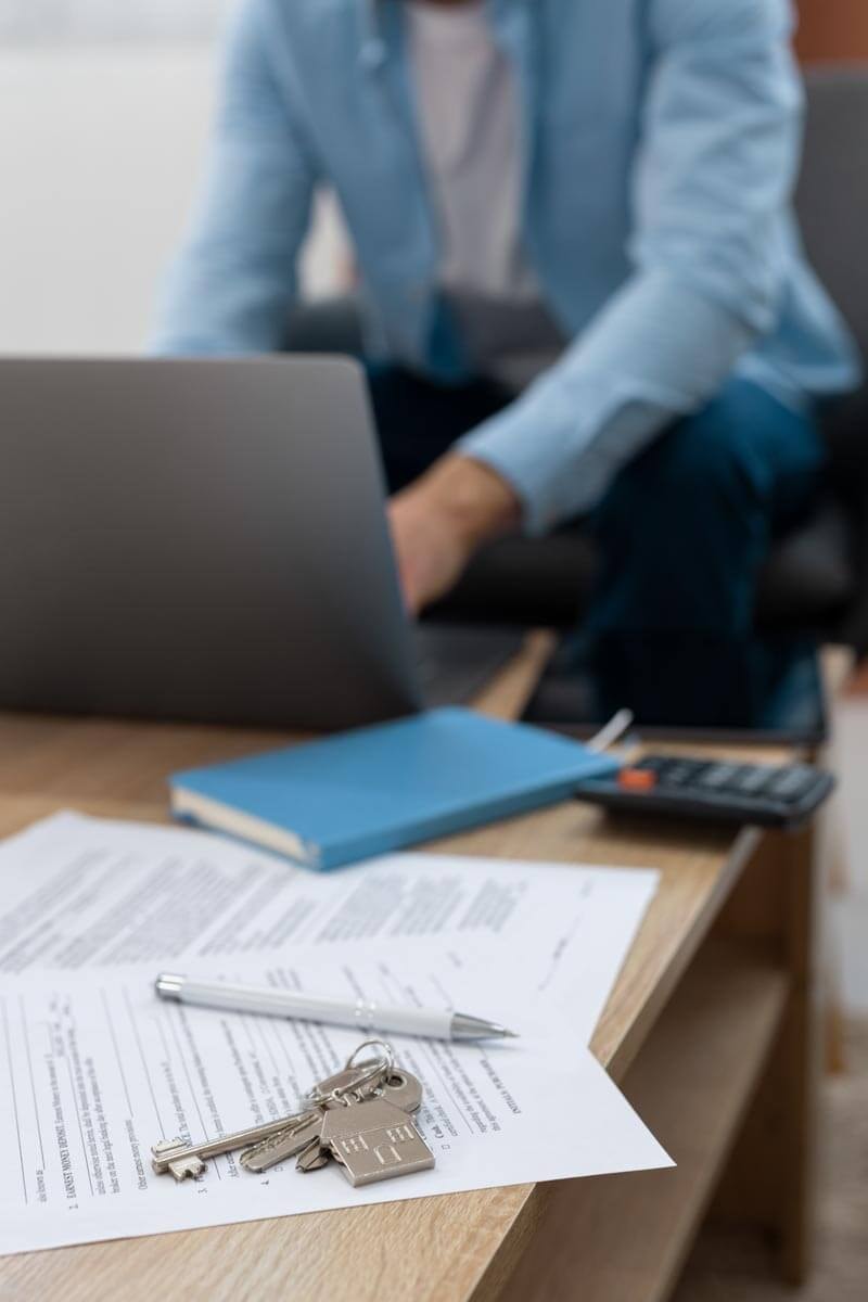 Close-up of a tenancy agreement lying on a table with a ballpoint pen and house keys beside it, while a person works on a laptop in the background.