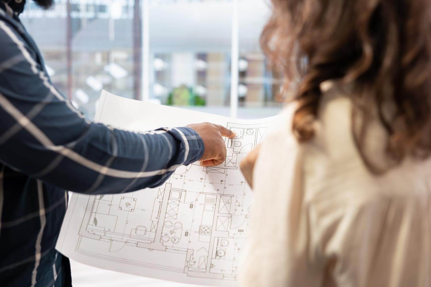 Rear view: a man shows a woman a bedroom on a black-and-white floor plan, which they hold up together