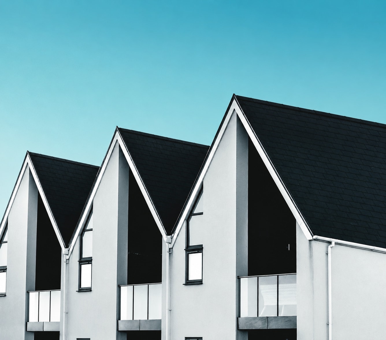 Modern terraced house with recessed balconies featuring glass panel railings set into the geometrically designed facade, and a steep pitched roof with black shingles against a blue sky.
