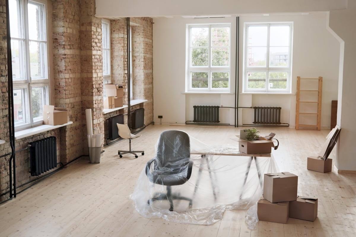 An empty loft-like space with exposed brick walls and large windows; in the centre stands an office chair wrapped in plastic film, surrounded by moving boxes, with an empty shelving unit against the wall.