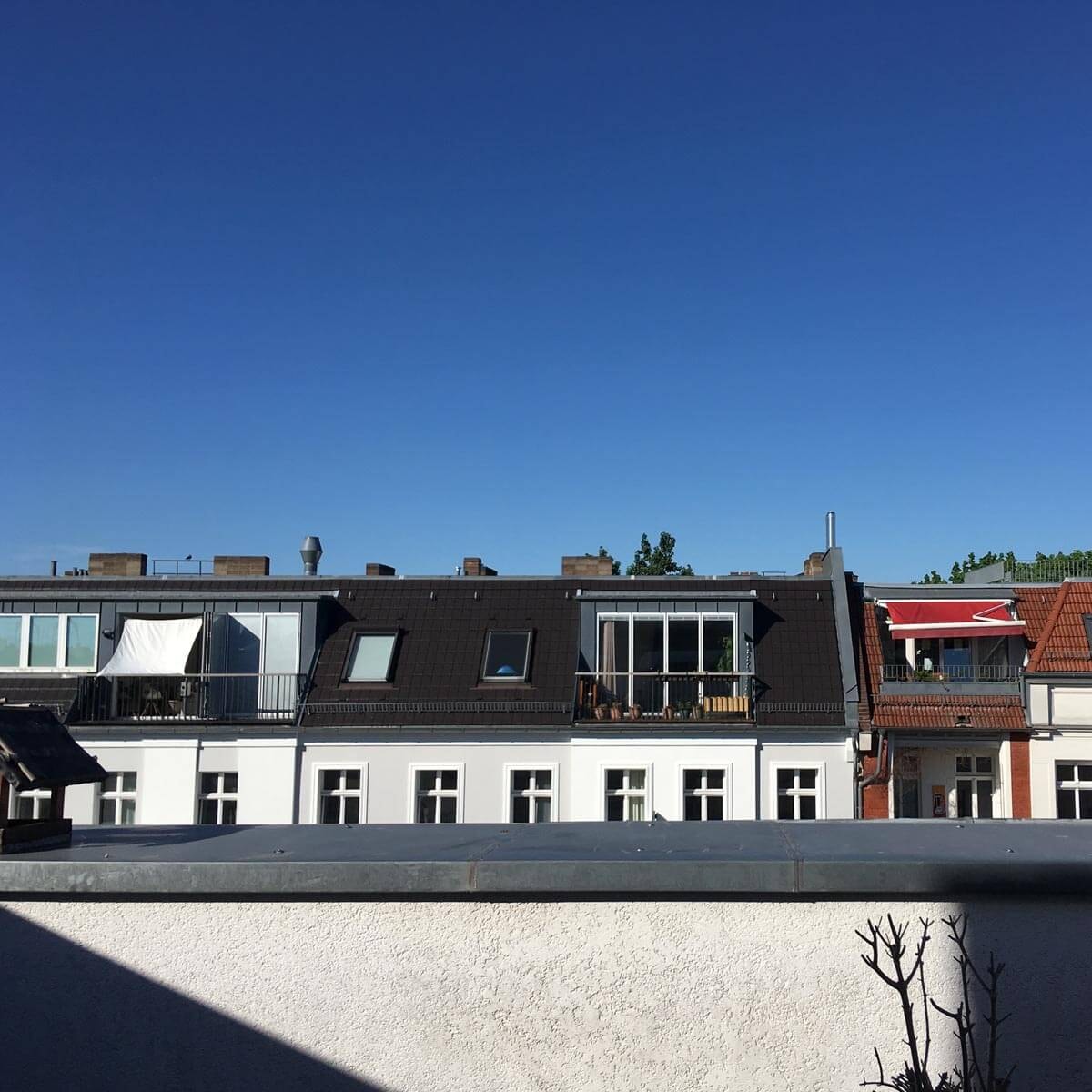 View from a roof terrace across a row of residential buildings with converted attics, dormers, and balconies against a bright blue sky; a grey parapet in the foreground.