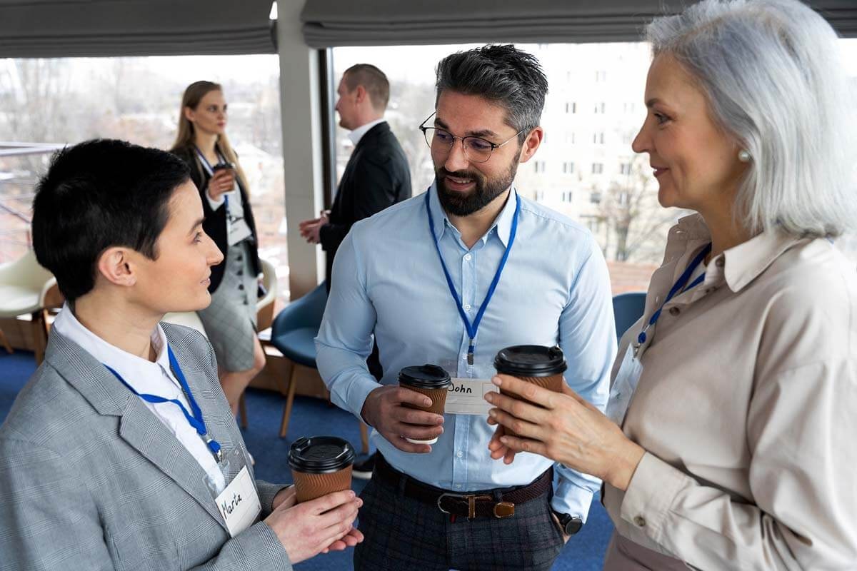 Three people holding coffee cups in conversation in the foreground, with two further people talking in the background in front of a wide window overlooking surrounding buildings.
