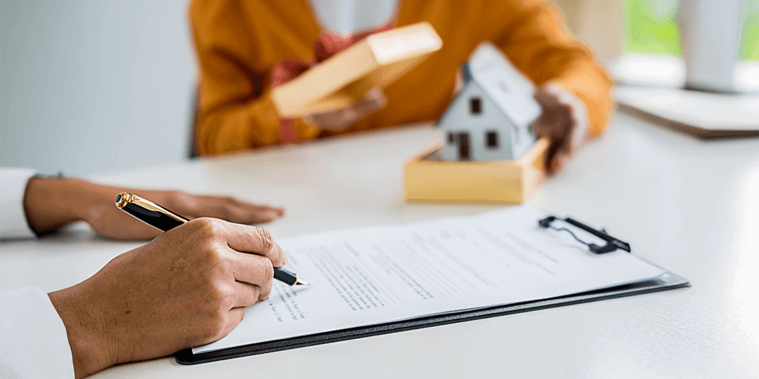 Close-up of a person at a table signing a document, while another person sitting further back slides a miniature house towards them.
