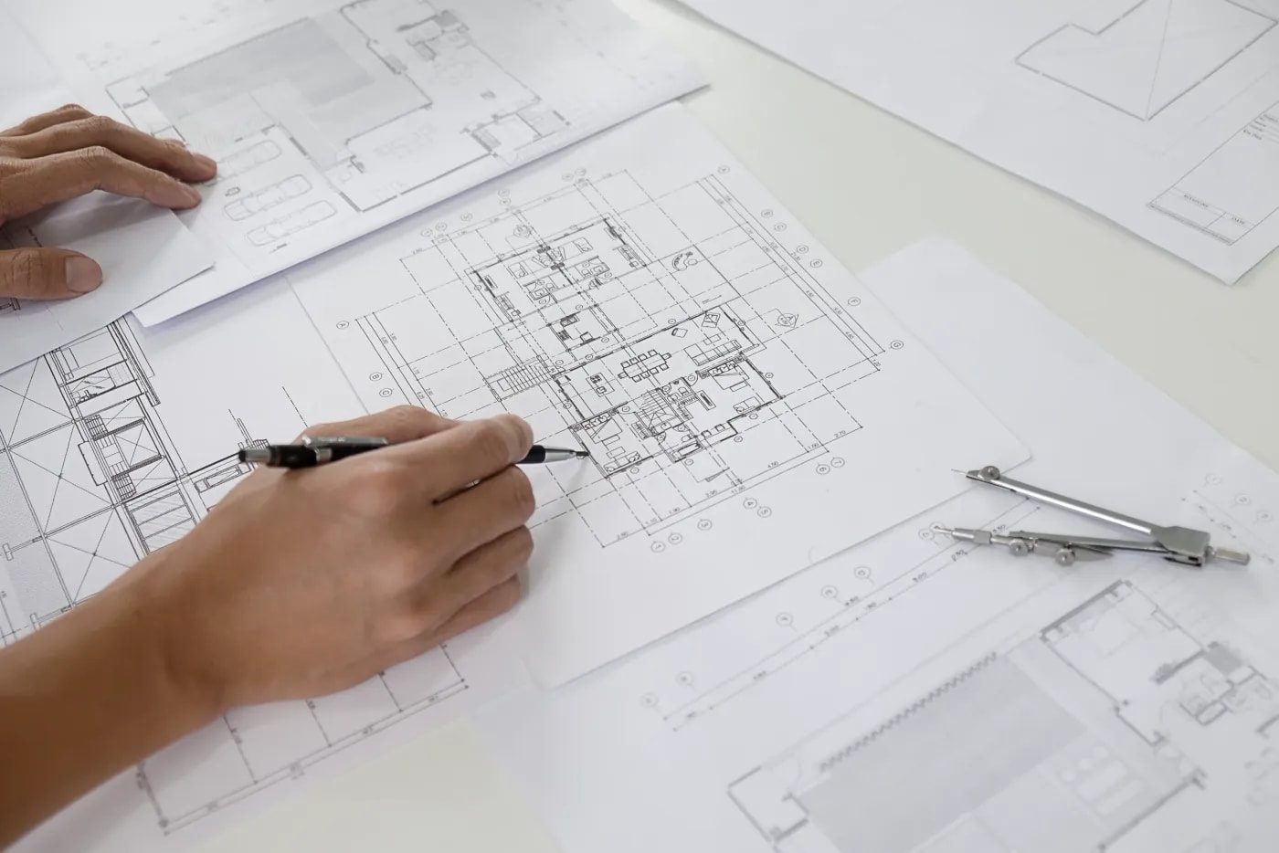 Close-up of a construction plan lying on a table among several other plans, with two hands, one of which holds a pen and traces over a detail on the plan
