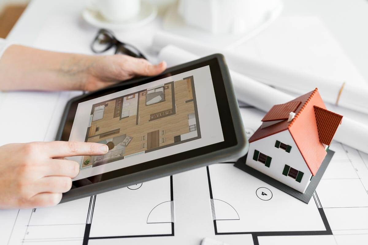 A person sits at a table with a floor plan spread out on it, holding a tablet showing a modern floor plan, with a small house miniature in front of them, while their hand points to a detail on the tablet