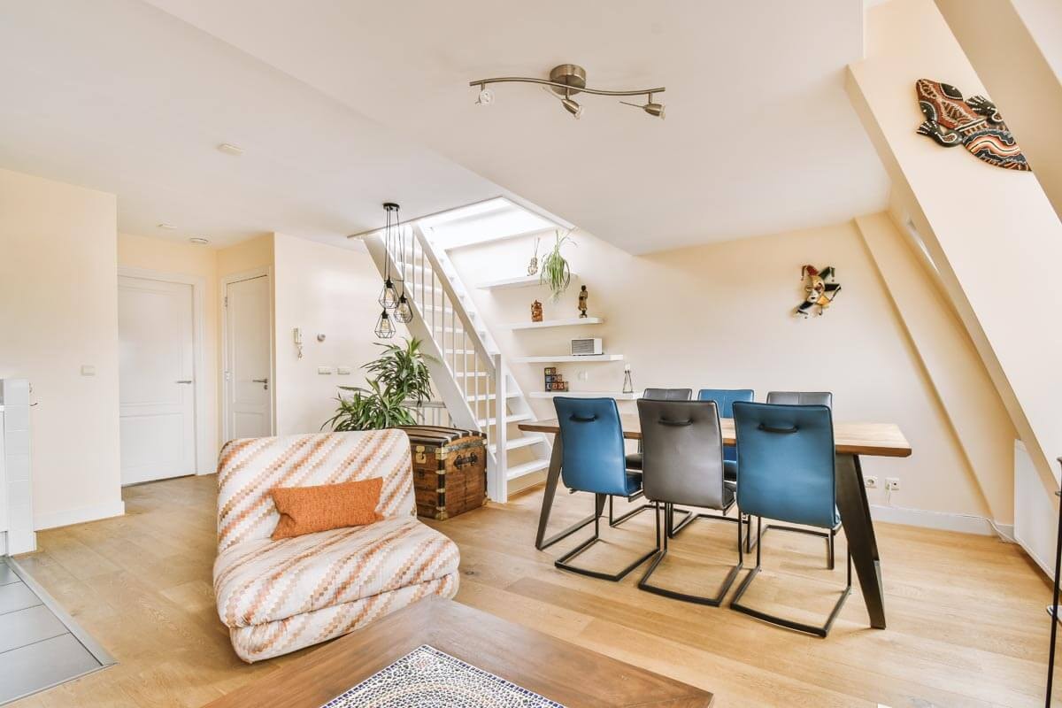 A mansard apartment with light parquet flooring and a characteristic sloped ceiling on the right; a dining area with a blue leather chair and wooden table, a patterned armchair with an orange cushion on the left, and a white wooden staircase leading upstairs in the background.