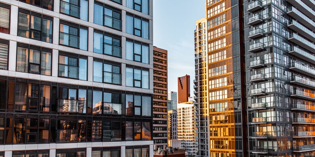 View between modern high-rise buildings with glass and balcony facades in a city, lit from the side by the evening sun.
