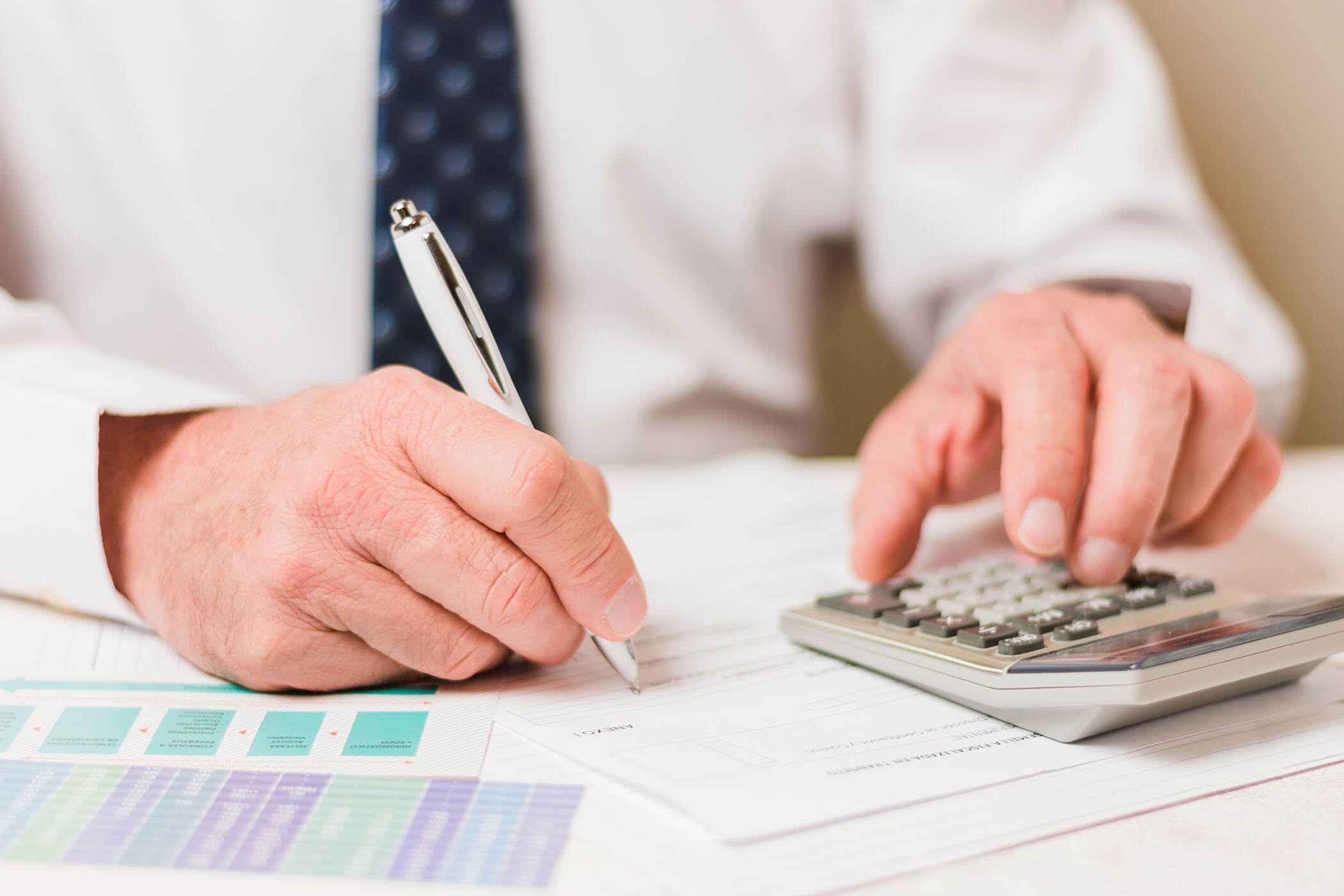 A person sits at a table with many documents, handwriting something on a form, apparently reading it off a calculator