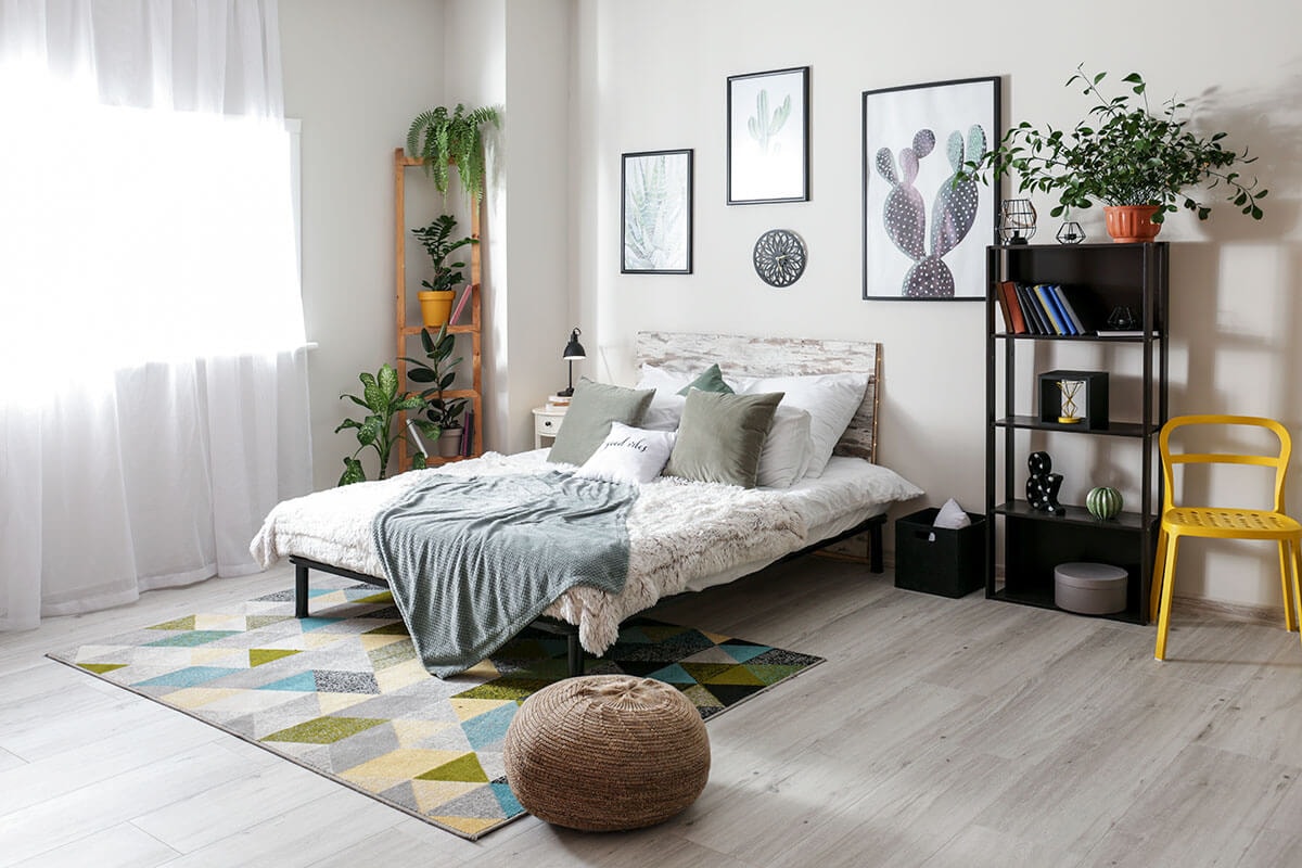 View from the corner of a room into a bedroom with a floor-length curtain in front of the window on the left, a futon bed in the centre beneath some cactus prints on the wall, and an open black shelf with a yellow chair.