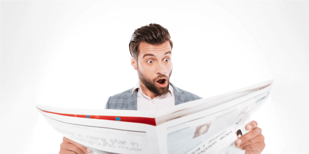 A man with an exaggeratedly astonished facial expression reads something in a real estate newspaper that he holds up in front of him with both hands