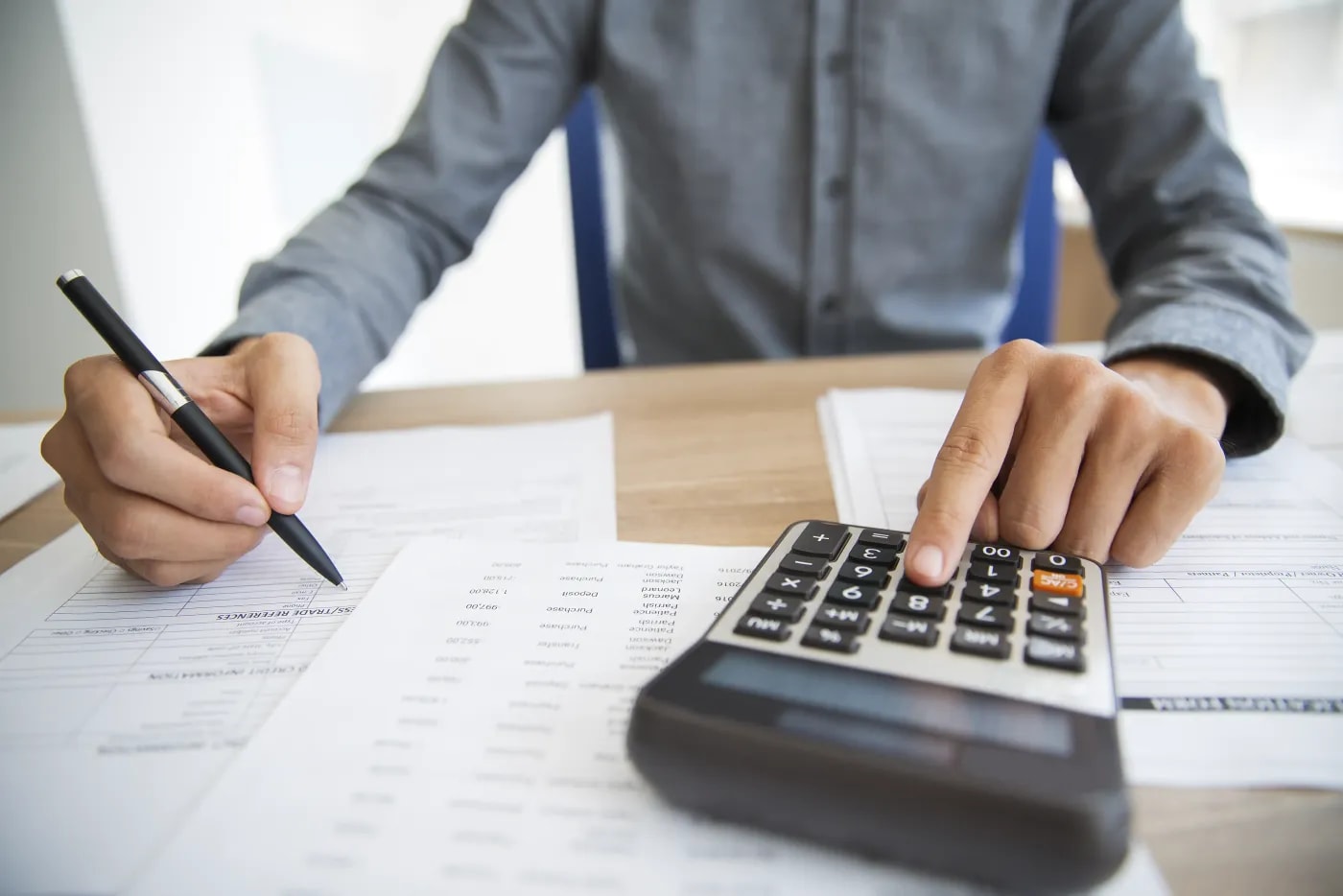 Front view of a person sitting at a desk, with a close-up of tabular documents from which the person is entering something into a calculator