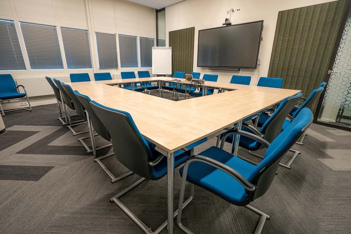 Four tables arranged in a circle with chairs around the outside; the windows in the background are fitted with blinds and a large monitor hangs on the wall beside the arrangement.