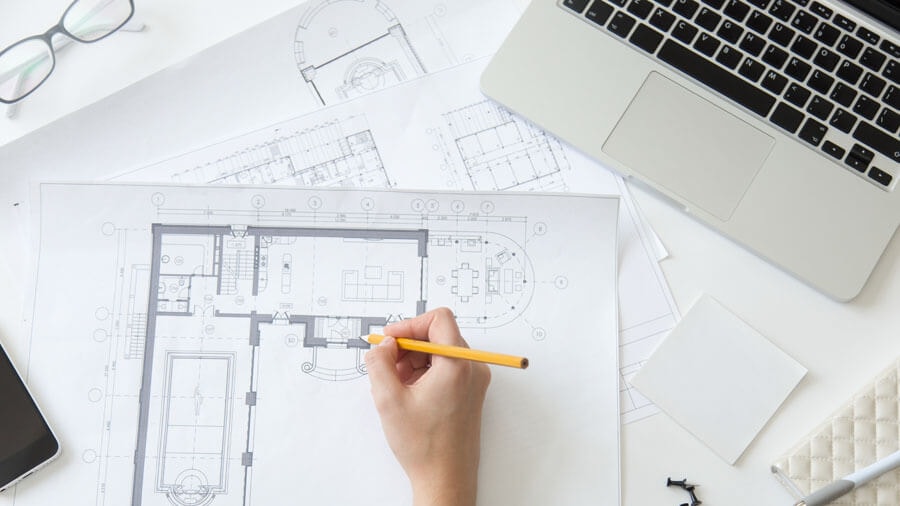 Top view of a desk with an open laptop and several documents, and a hand noting something on a floor plan with a pencil.
