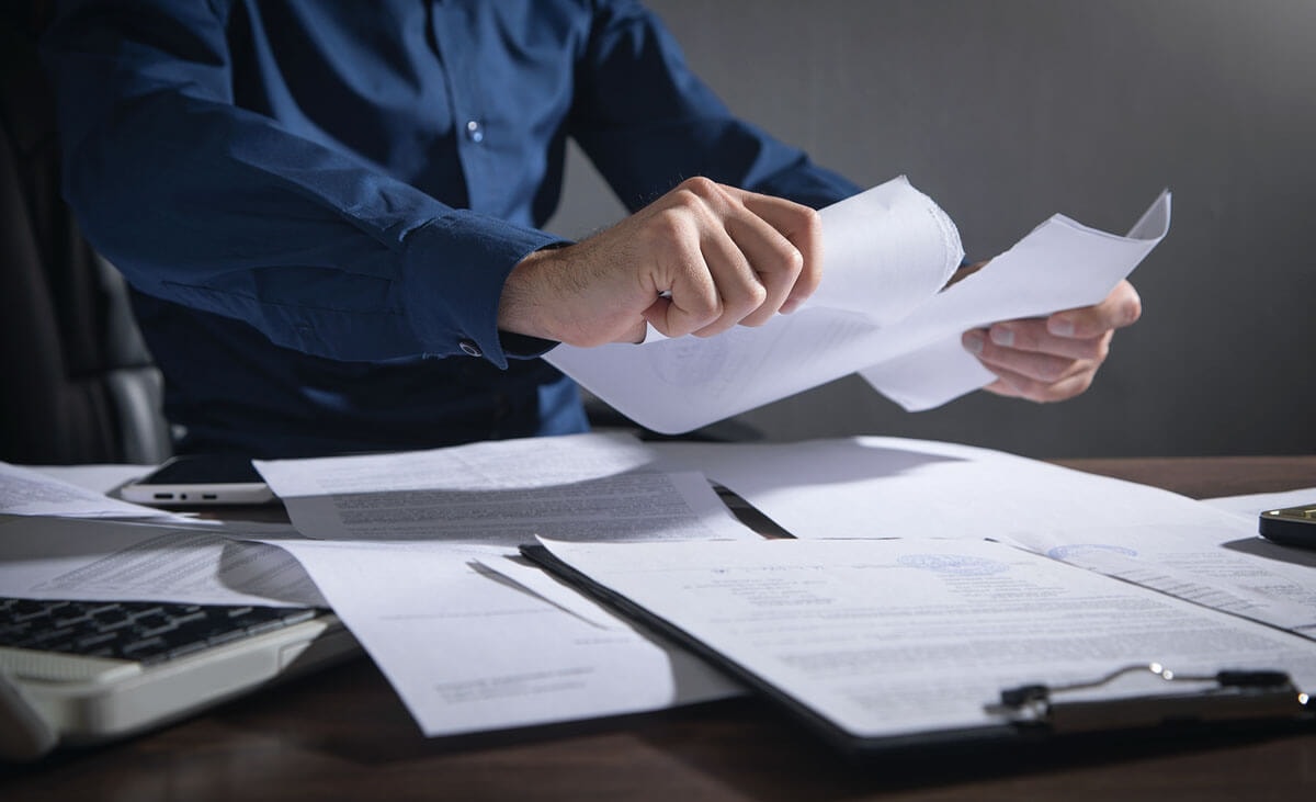 A man in a blue shirt going through various documents spread out on the table in front of him.