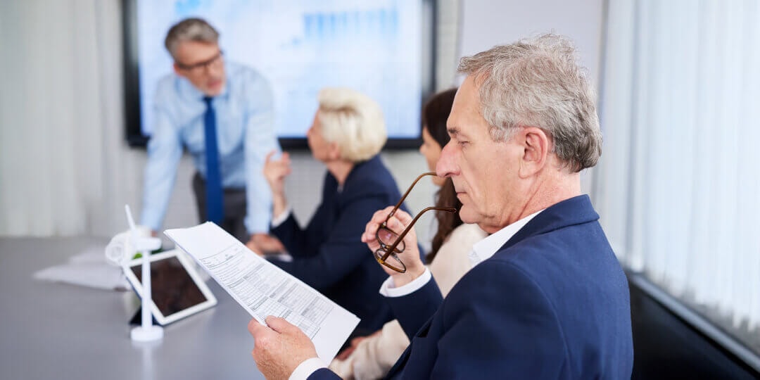 Older businessman concentratedly reading a document, with a team presentation on a screen in the background.
