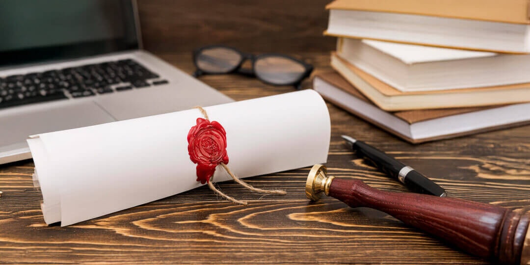 Rolled certificate with a red wax seal on a wooden table, alongside a stack of books, glasses, a laptop and a seal stamp.