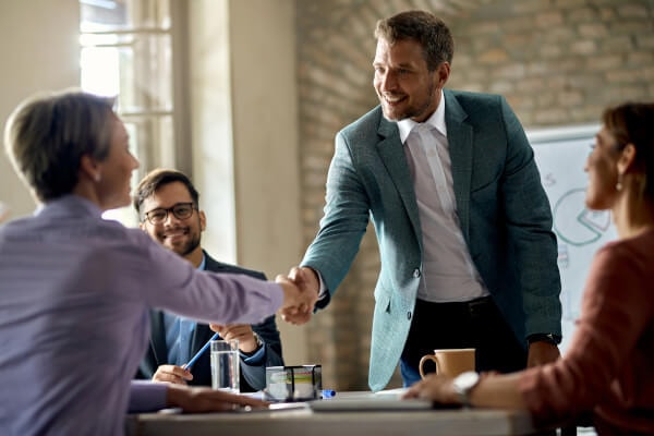 Business partners are visible at a meeting table full of documents, two of whom are smiling and shaking hands