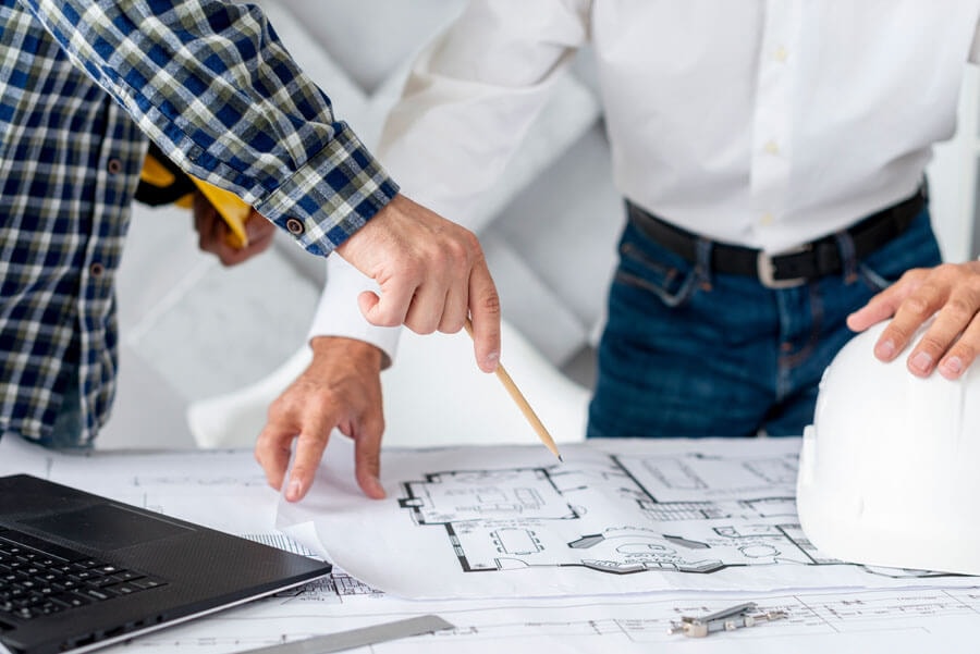 Three men's hands are engaged with a floor plan: one hand rests on a white hard hat, one holds a pencil and points to a detail, and the third hand holds down a corner of the plan