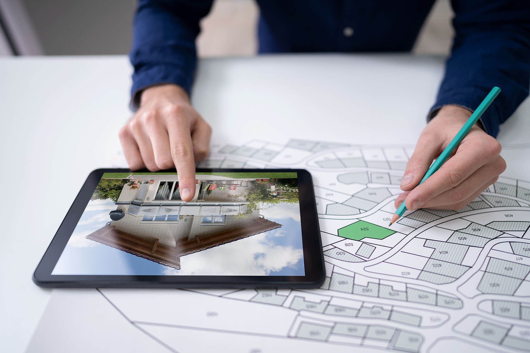 Nahansicht eines Flurplanes auf dem ein Tablet liegt, das ein zweigeschossiges Einfamilienhaus zeigt und eine Person, die mit einer Hand ein Grundstück markiert und mit der anderen auf das Haus zeigt.