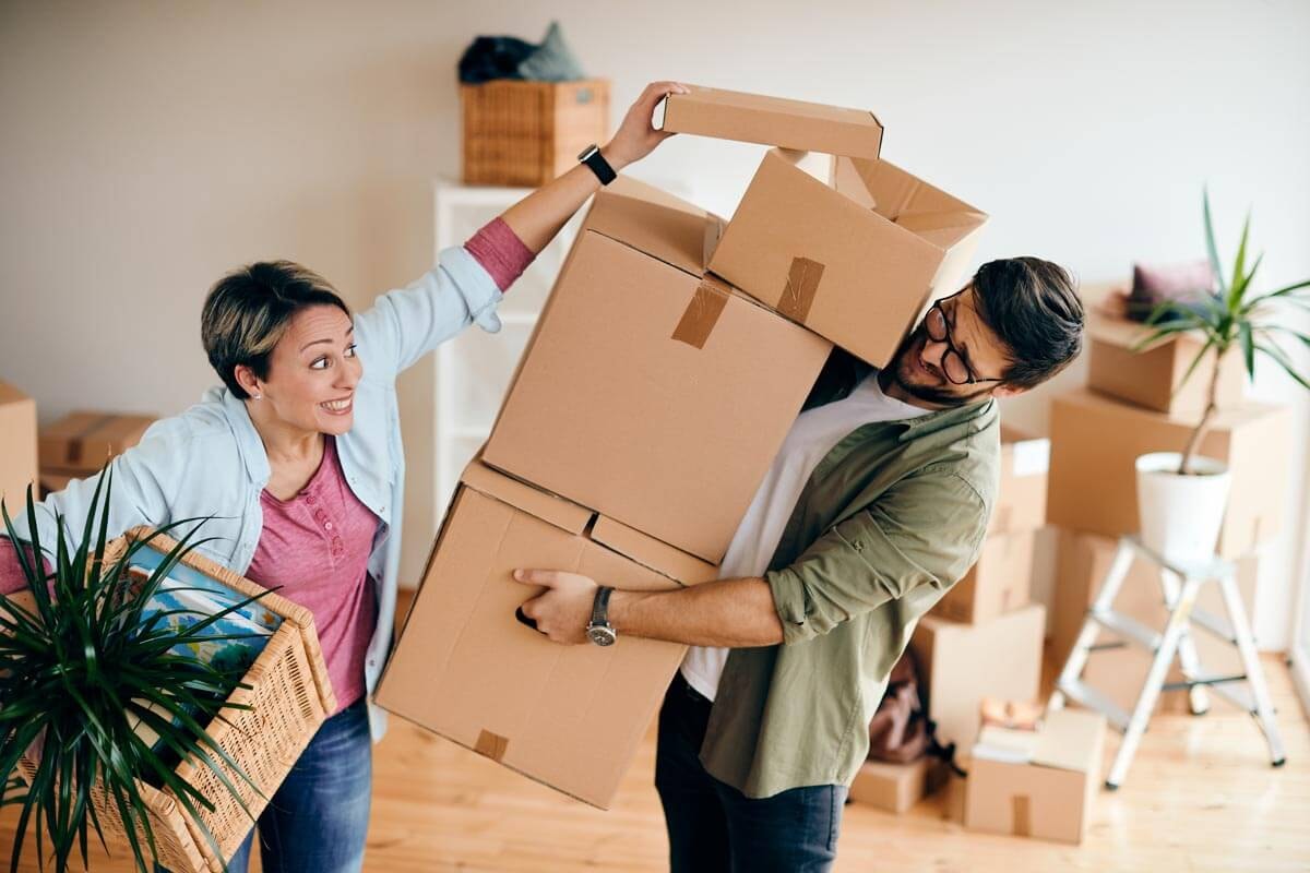 A couple during a move: the man is balancing a tipping stack of moving boxes while the woman laughs and reaches for the top box; she is also carrying a basket with a plant.