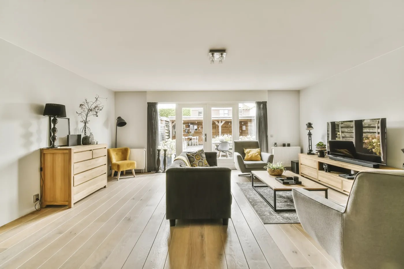 Bright, elongated living room with wooden flooring and patio doors at the far end, through which a garden with a garden shed is visible.