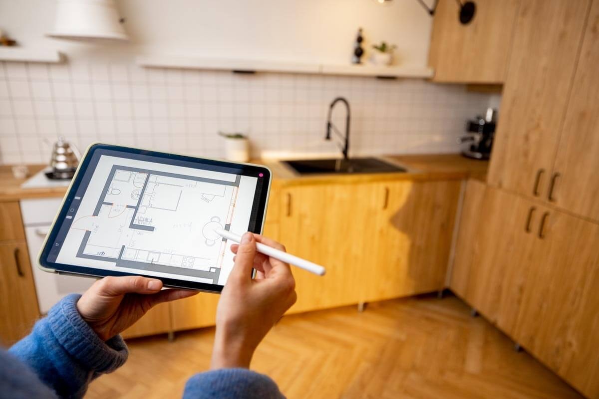 A person holds up a tablet in a kitchen with pine wood cabinets and parquet flooring.
