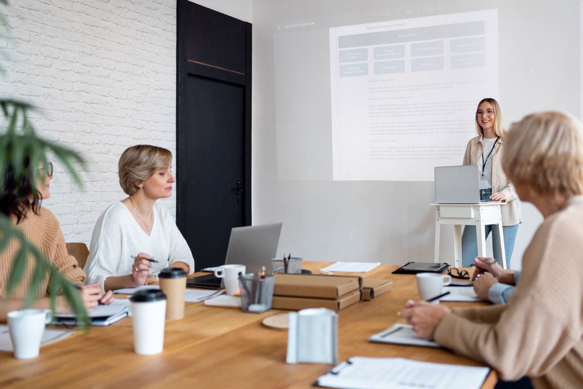 Several women seated at a cluttered wooden table during a meeting, with one woman giving a PowerPoint presentation.