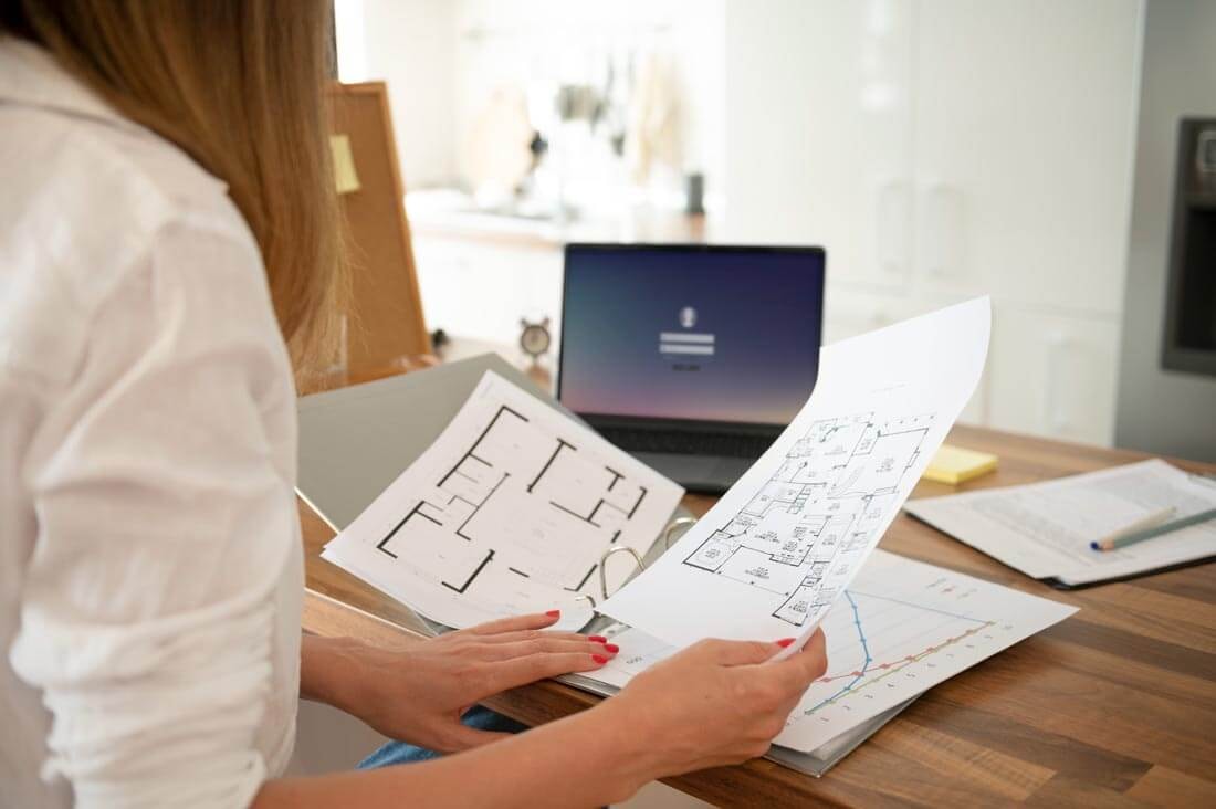 View past a woman's arm to a binder containing various construction documents, from which she is removing a floor plan to compare it with another floor plan lying beside it.
