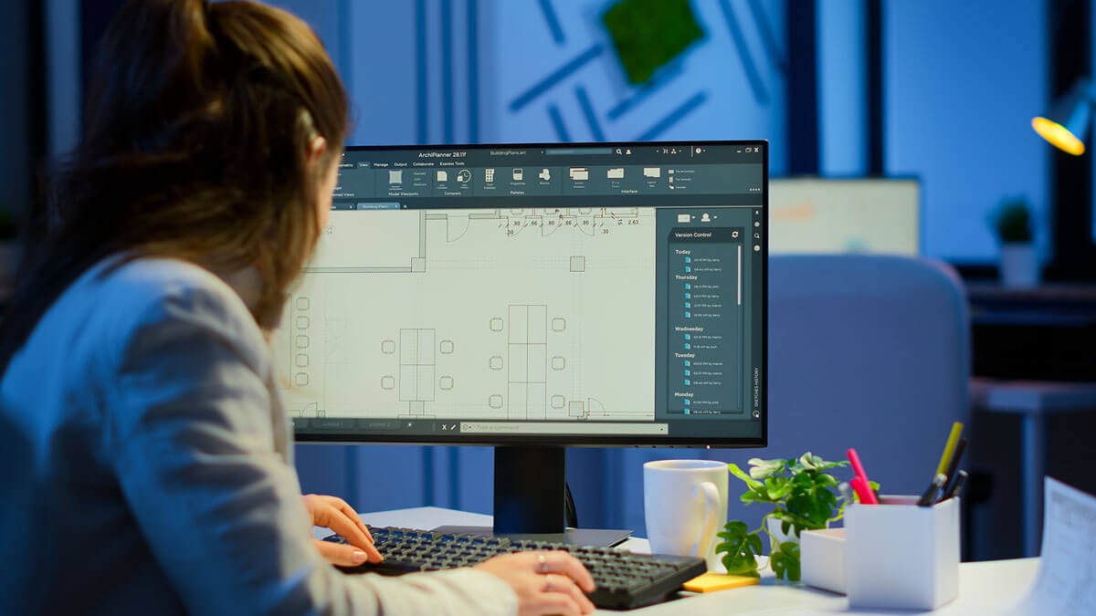 Rear view of a woman sitting at a desk with a PC in an office, hands on keyboard and mouse, editing a floor plan visible on the monitor.