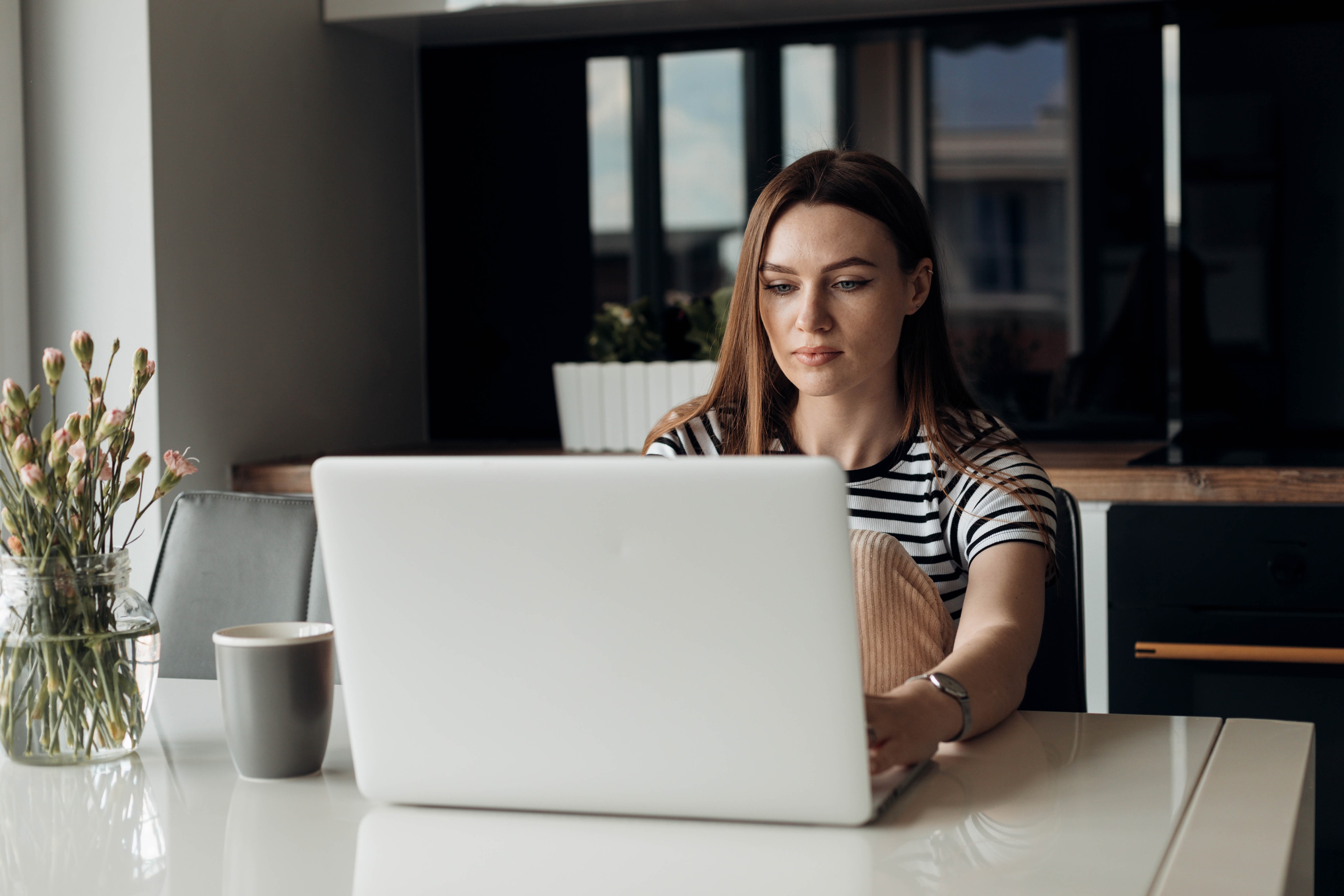 A woman sits at a kitchen table working on a laptop, with dark kitchen cabinets visible in the background.