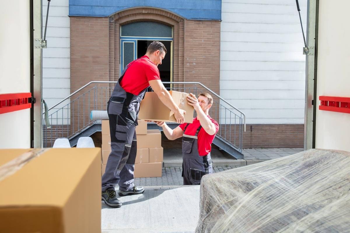 Two removal workers in red workwear pass boxes to each other from the open loading area of a removal van; in the background a brick building with a staircase entrance.