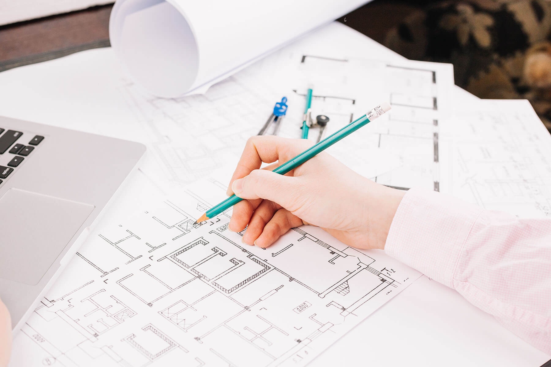 A hand makes pencil notes on a floor plan, with a laptop beside it.