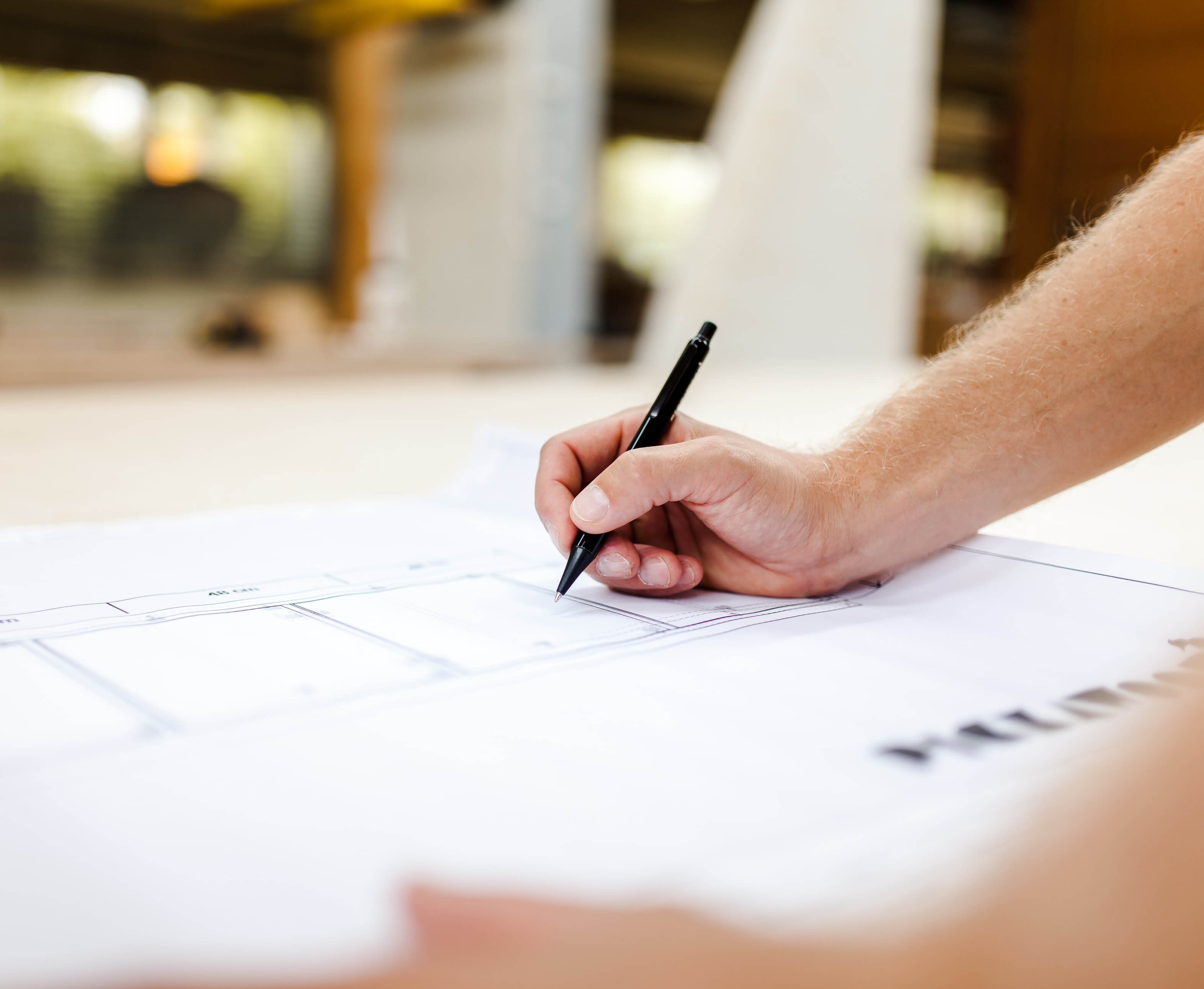 Close-up of a hand entering lines into a large-format floor plan with a black pen, with an office visible in the background.