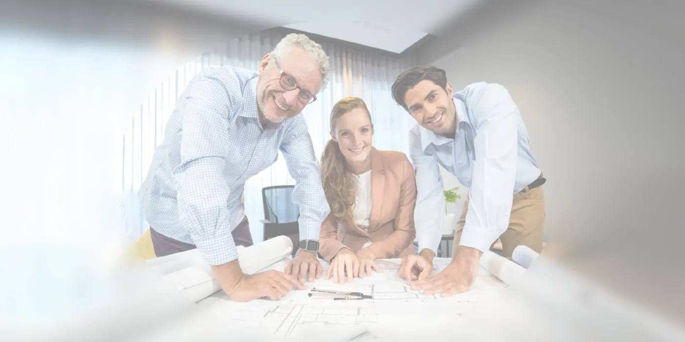 Two men and a woman lean over a floor plan on the table in front of them, propped up on their hands or arms, smiling warmly at the camera